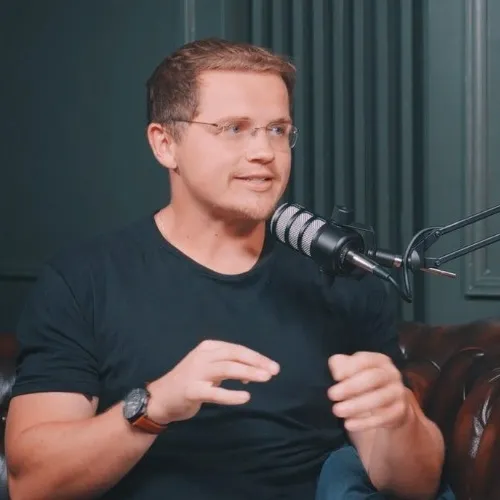 Man with glasses speaking into a microphone while gesturing with his hands, seated on a brown leather couch.